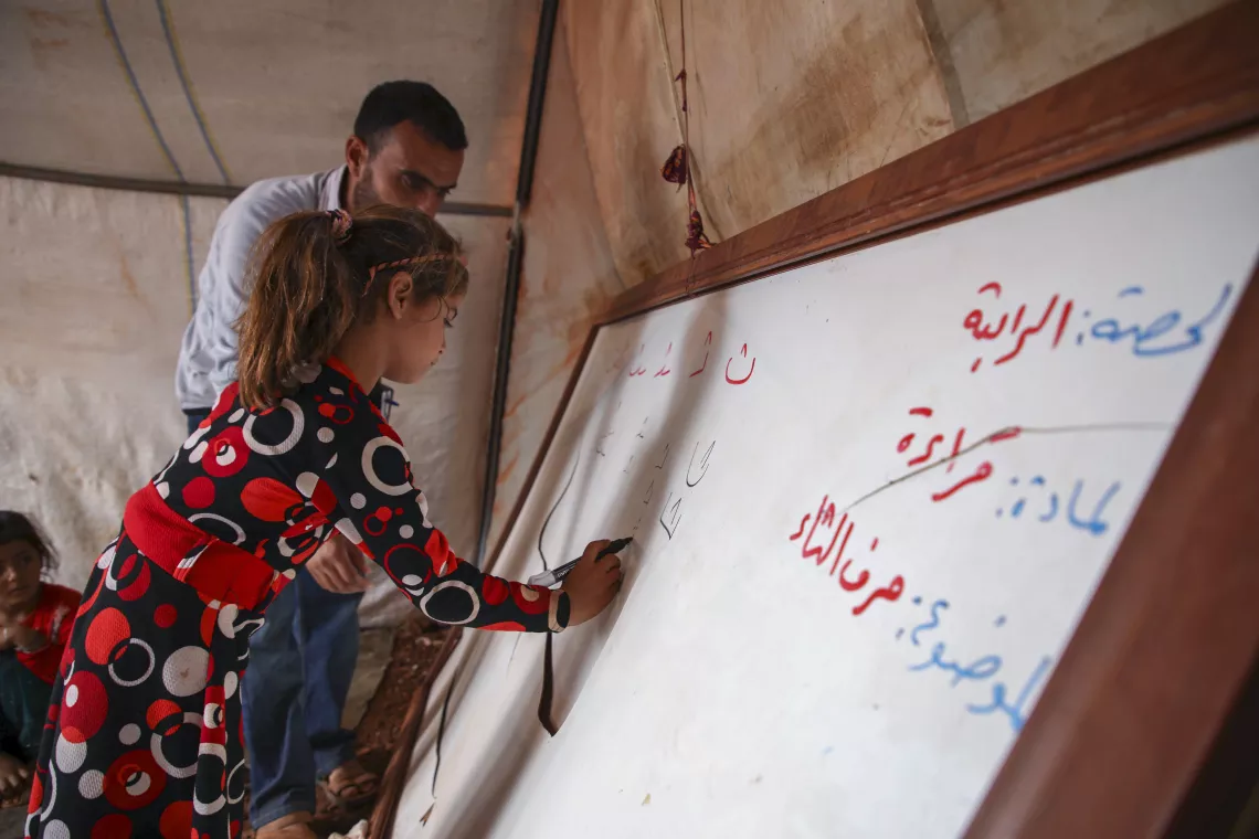 A young girl is writing in Arabic on a white board, supported by her male professor