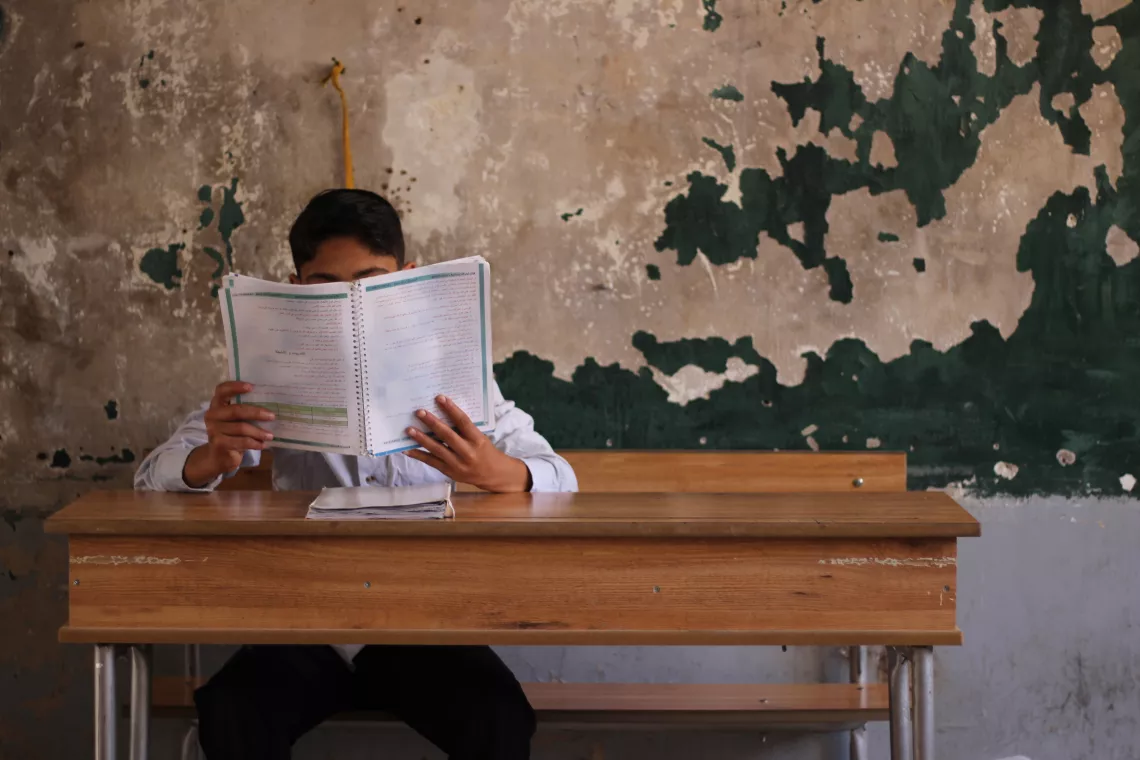 A young boy is reading a book in Arabic, sitting in a classroom with decrepit w